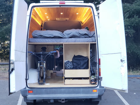 White RV rental van's open rear reveals a custom wood interior with bed, bike storage, helmet, & gear. Warm lights glow.