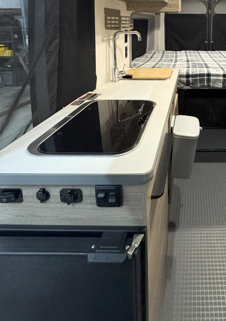 Modern RV rental interior: kitchenette with sink, induction cooktop, and sleeping quarters in the background.