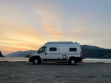 Experience an RV rental adventure! Silver van parked by a scenic lake with mountains at sunset.