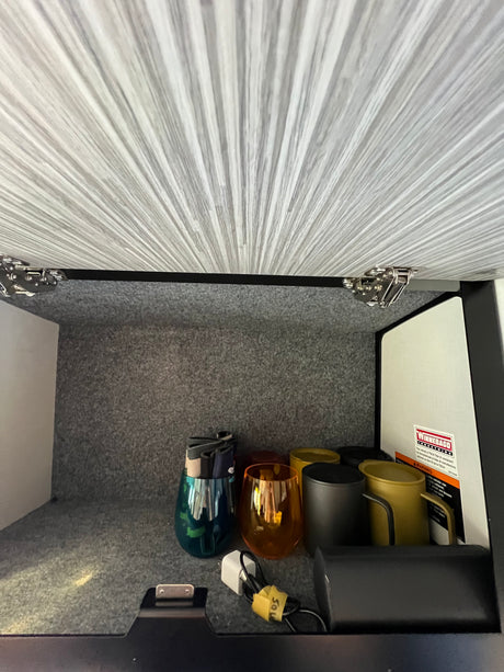 Inside an RV rental overhead compartment with textured gray walls, holding colorful cups, a charging cable, and a Winnebago label.