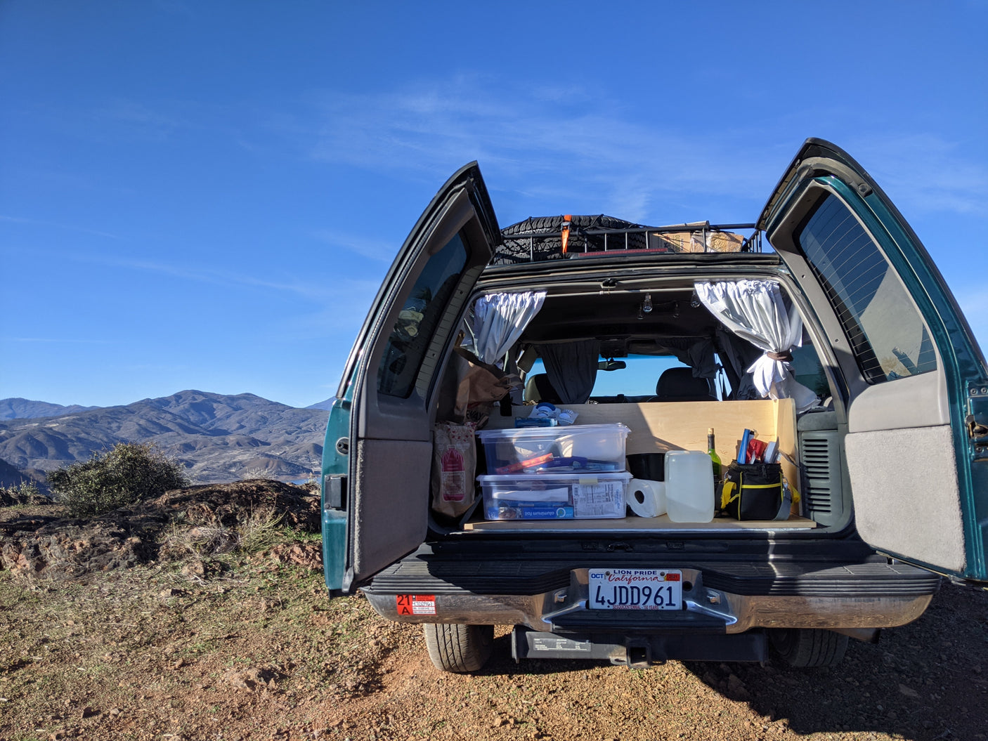 Rear of a converted SUV/van set up for rv rental car camping, showcasing organized gear, curtains, and mountain views.