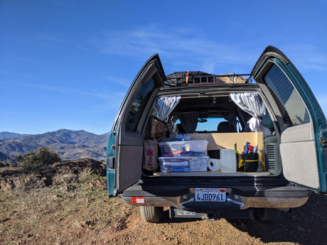 Rear of a converted SUV/van set up for rv rental car camping, showcasing organized gear, curtains, and mountain views.