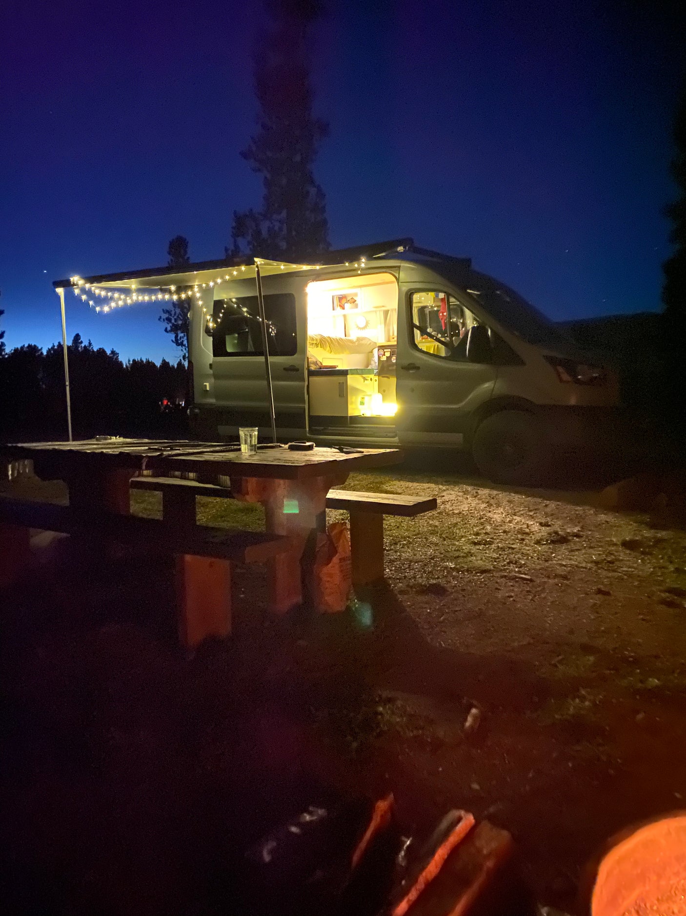 Experience a cozy night in this RV rental, with string lights illuminating the camper van and picnic table under a twilight sky.
