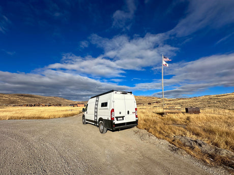 An RV rental (white camper van) at Bodie State Historic Park, a ghost town. Flags, dry landscape, and old buildings visible.