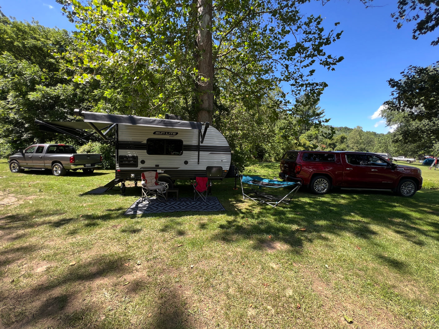 Relaxing RV rental setup at a shaded campsite with a travel trailer, chairs, and parked vehicles under green trees.