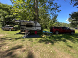 Relaxing RV rental setup at a shaded campsite with a travel trailer, chairs, and parked vehicles under green trees.