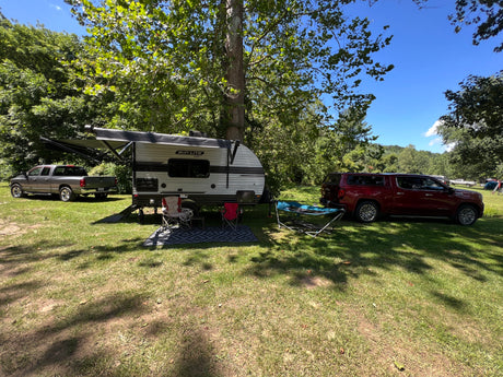 Relaxing RV rental setup at a shaded campsite with a travel trailer, chairs, and parked vehicles under green trees.