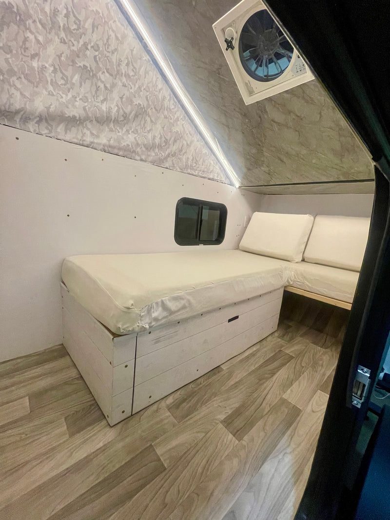 Cozy RV rental interior featuring a white mattress bed with storage, L-shaped seating, small window, and wood-look floor. — photo 7