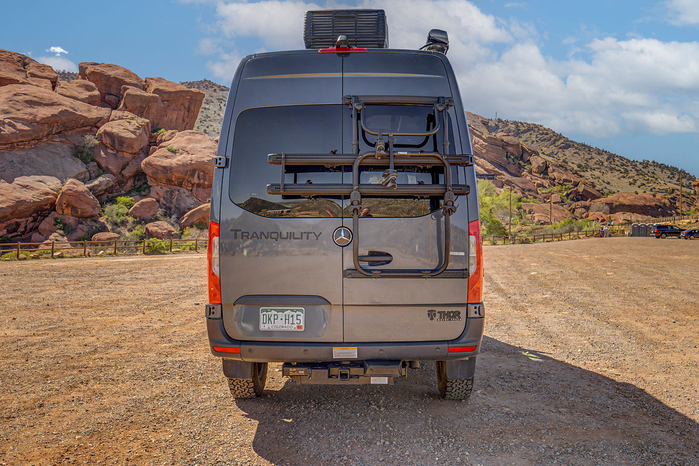 Rear of a gray rv rental camper van with a bike rack, parked in a scenic red rock mountain landscape under a blue sky.