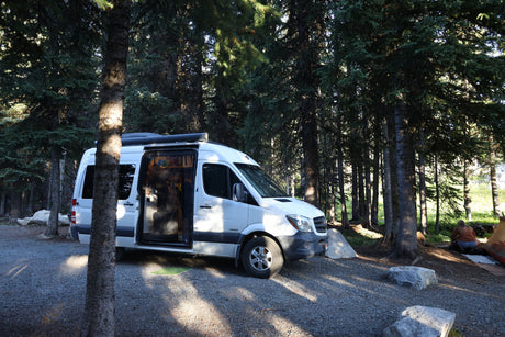 White rv rental parked on gravel in a dense forest, side door open, revealing interior. A person crouches by a tent on the right.