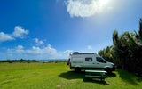 Perfect RV rental spot: silver camper van on green grass by the ocean, with a picnic table & sunny sky.