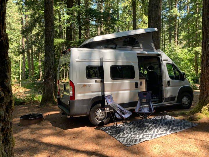 Enjoying nature! A silver camper van RV rental parked in a sunny forest campsite with chairs, rug, and fire pit.