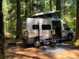 Enjoying nature! A silver camper van RV rental parked in a sunny forest campsite with chairs, rug, and fire pit.