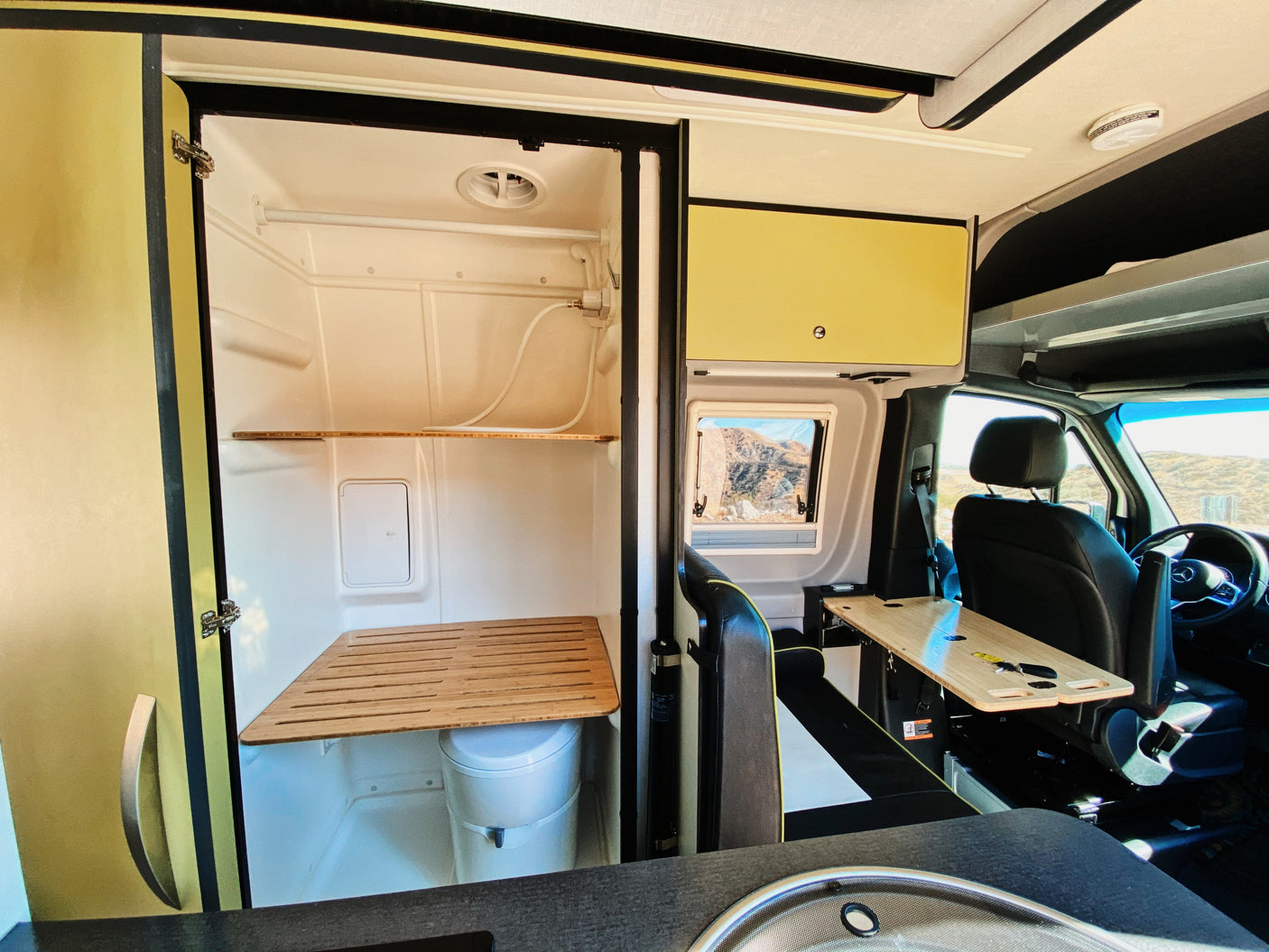 Compact RV rental interior featuring a shower/toilet, kitchen sink, driver's cabin, and scenic window view.