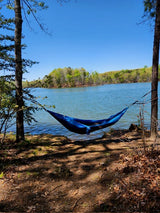 Escape with an RV rental! A blue hammock overlooks a calm lake and distant green trees under a clear sky. Relaxing view.