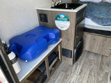 Compact RV rental interior featuring a sink, control panel, bed, and blue water storage.