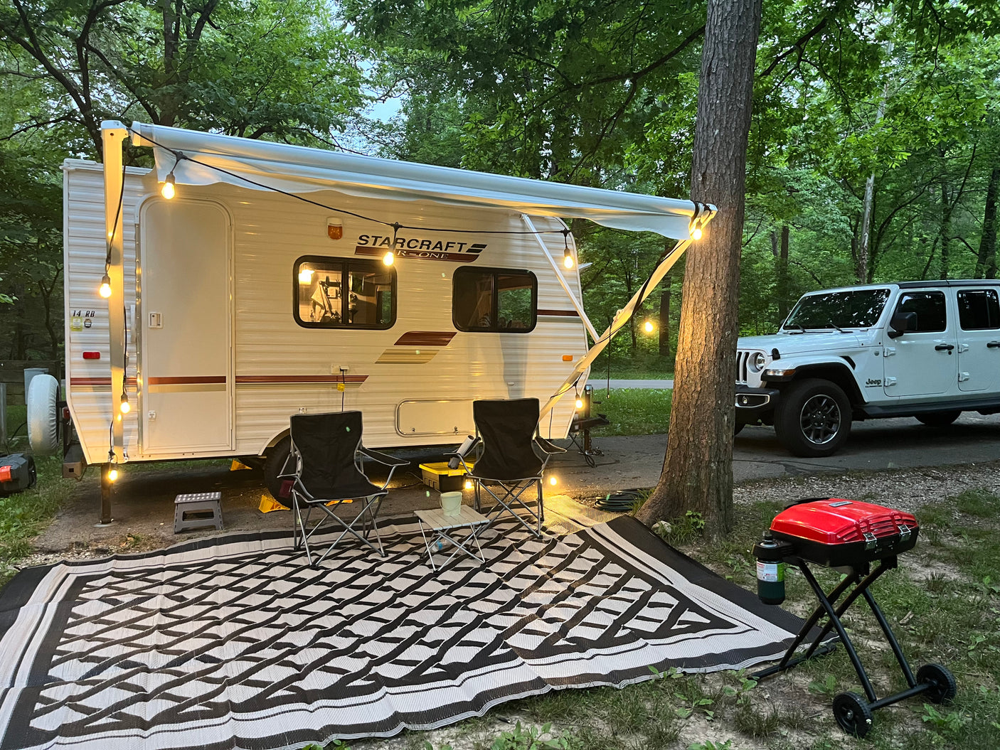 Cozy RV rental campsite setup with string lights, chairs, outdoor rug, and a grill next to a white Jeep among trees.