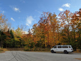 An RV rental, a white van, parked in a vibrant autumn forest with colorful trees under a blue sky.
