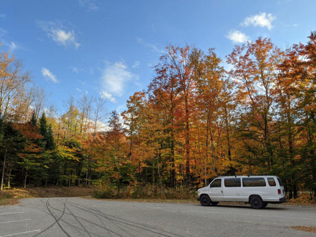 An RV rental, a white van, parked in a vibrant autumn forest with colorful trees under a blue sky.