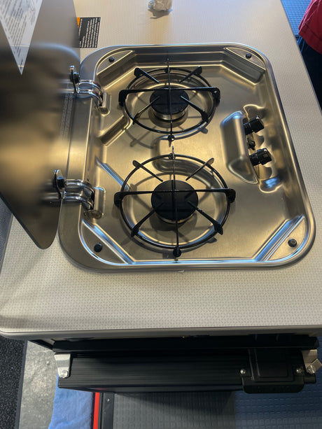 RV rental: A stainless steel, two-burner RV stove with its protective lid open, revealing black grates and control knobs.