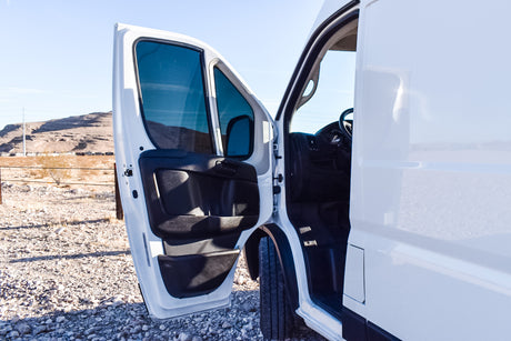 This white RV rental van with an open driver's door shows its interior, parked on a gravel lot with mountains.