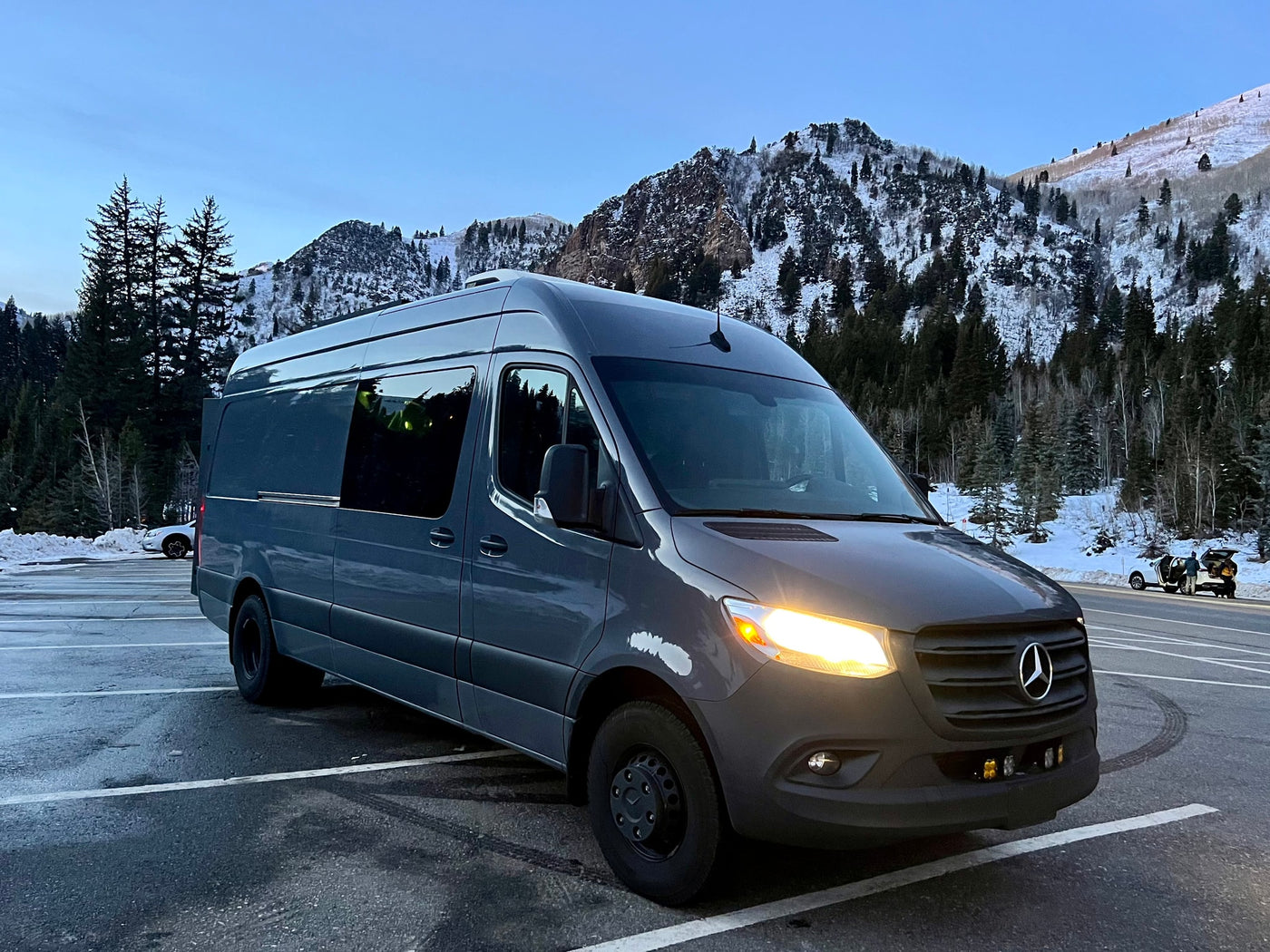 A dark gray Mercedes Sprinter camper van, likely an RV rental, parked in a snowy mountain landscape at dusk with headlights on. — photo 3
