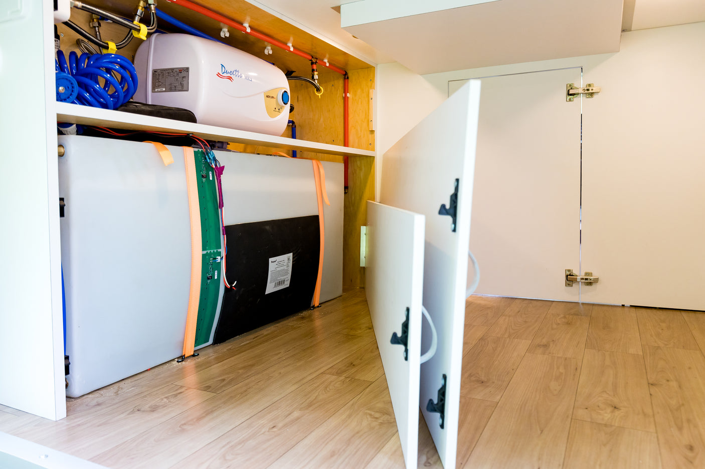 An RV rental's utility compartment reveals a white water heater, clear water tanks, and blue/red plumbing lines.