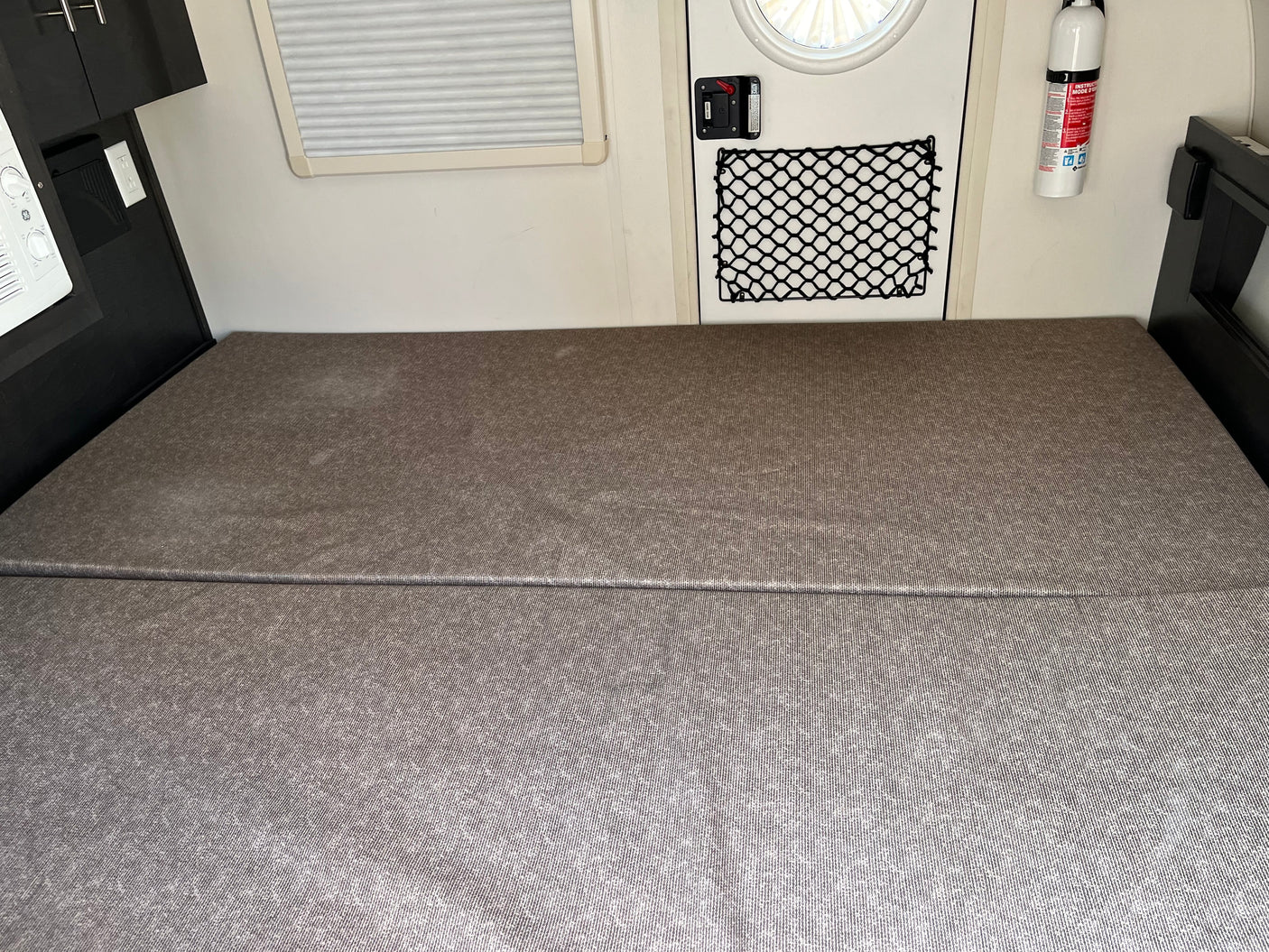 RV rental interior: a gray modular bed, adjacent cabinets, door with storage net, and fire extinguisher.
