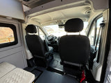 Spacious RV rental interior with black front seats and dashboard. Left shows cabinet storage and bed, with rocky outdoor views.