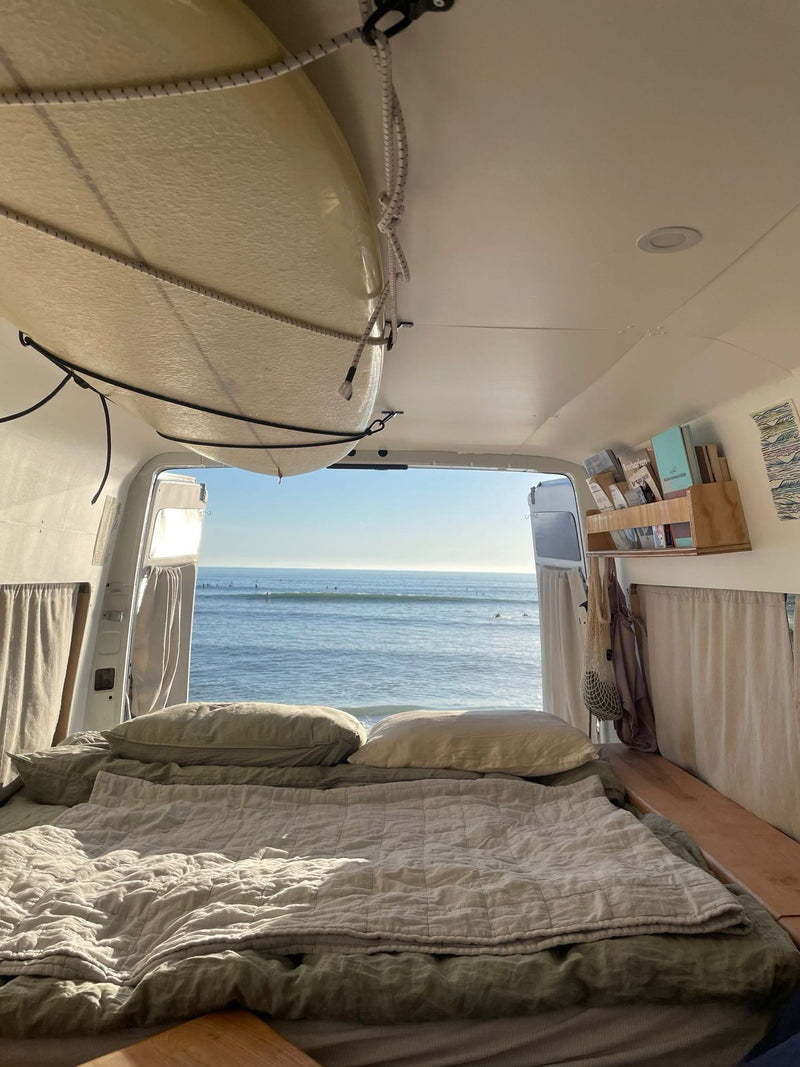  parked street-side in golden hour light.|A white Dodge Sprinter van (RV rental) parked on a residential street. Reflections of sky/poles in rear windows. Maine plates visible.|Inside an RV rental: a cozy bed faces an ocean view with surfers. A surfboard is stored above.|Ocean vista from an RV rental van — photo 7
