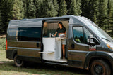 An RV rental experience: Woman inside a grey camper van, organizing supplies in the kitchen area, surrounded by a lush forest.