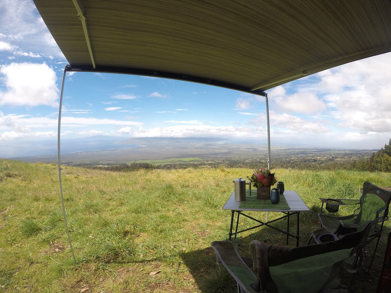 This RV rental setup features an awning shading a table with coffee and flowers, two chairs, and a stunning view of mountains & ocean. — photo 10