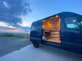 Discover adventure with our RV rental. See the cozy, lit wooden interior of this blue camper van at sunset outdoors.