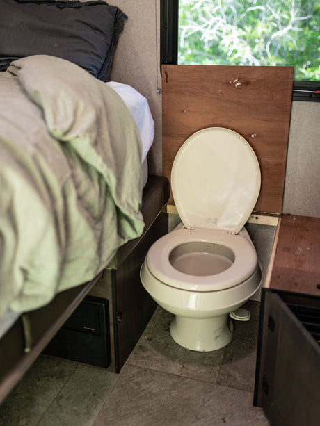 Inside an RV rental: a compact toilet with open lid, beside a bed and window with outdoor greenery.