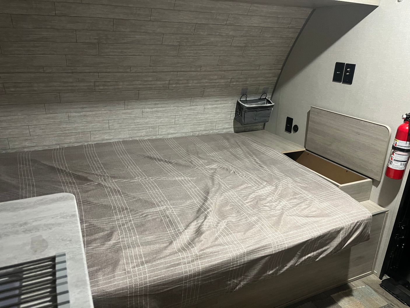 An RV rental bedroom featuring a plaid bed, integrated storage, and a fire extinguisher for safety and comfort.