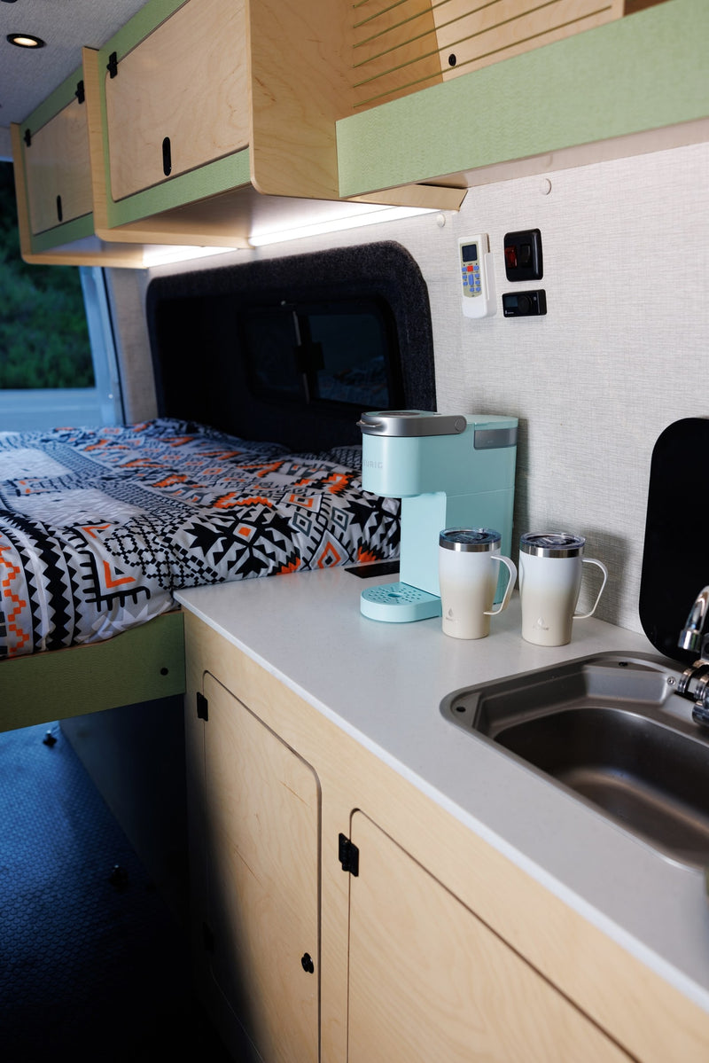 RV rental interior showcasing a cozy bed with patterned bedding, a compact kitchen with coffee maker, sink, and wooden storage cabinets. — photo 6