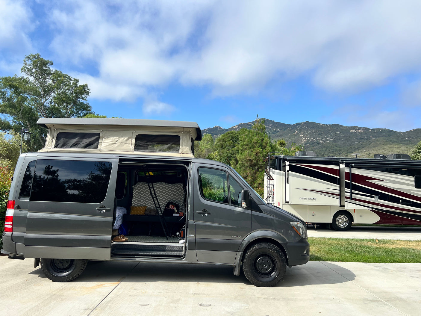 A gray pop-top camper van, possibly an RV rental, with its door open next to a large motorhome in an outdoor setting.