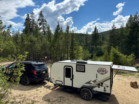 White Rubicon travel trailer (RV rental) towed by a black SUV, parked at a peaceful mountain forest campsite.