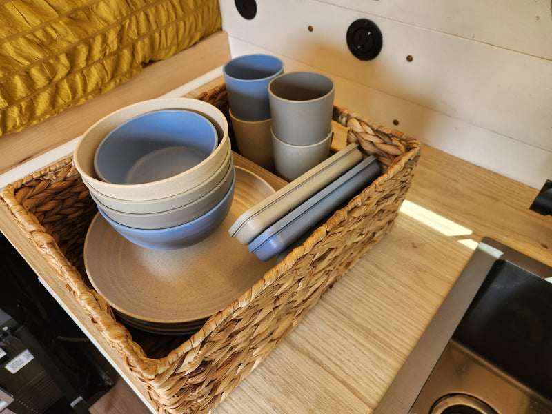 Organized RV rental kitchen with a woven basket holding stackable blue, gray, and beige dishes on a light wood counter beside a sink. — photo 15