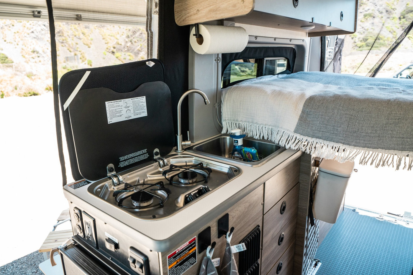 rv rental interior: A compact camper kitchen featuring a two-burner stove, sink with supplies, and a cozy bed in the background. — photo 12