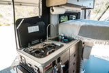 rv rental interior: A compact camper kitchen featuring a two-burner stove, sink with supplies, and a cozy bed in the background.