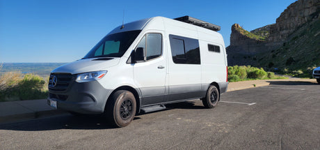 An RV rental, a white Mercedes Sprinter van, parked at a scenic viewpoint overlooking a valley and rock formations.
