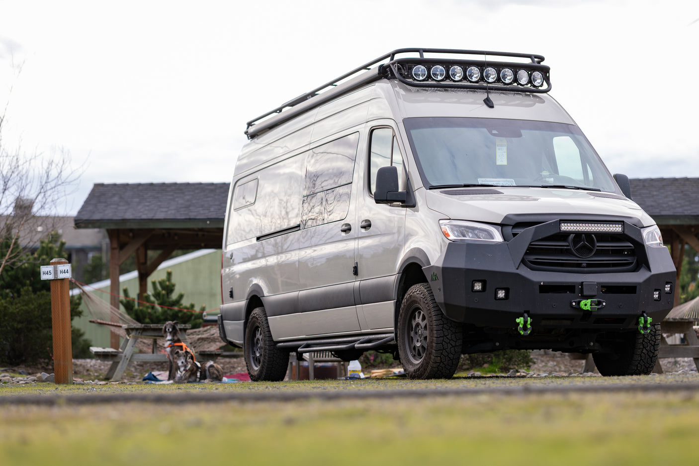 Rugged adventure RV rental van parked outdoors, featuring off-road gear, roof lights, and two dogs nearby.