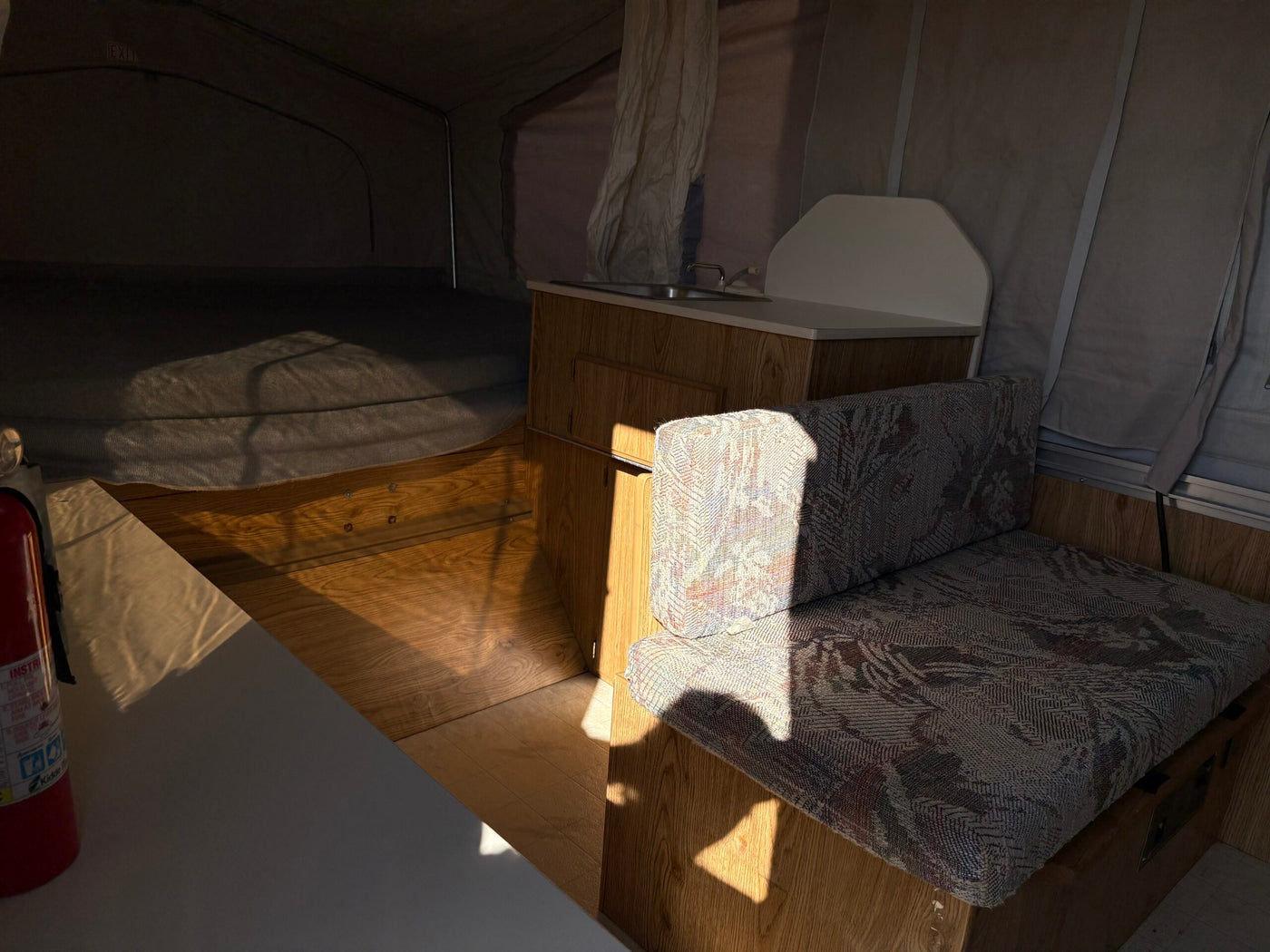 Inside a cozy RV rental pop-up camper: compact kitchen, bed, & dinette area with patterned cushions. Sunlit interior. — photo 8