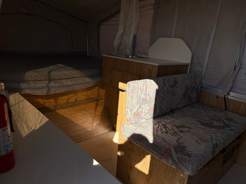 Inside a cozy RV rental pop-up camper: compact kitchen, bed, & dinette area with patterned cushions. Sunlit interior. — photo 8