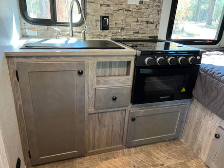 Modern RV rental kitchen featuring a sink, GreyStone stove with oven, and stylish wood-look cabinets. Scenic window view.