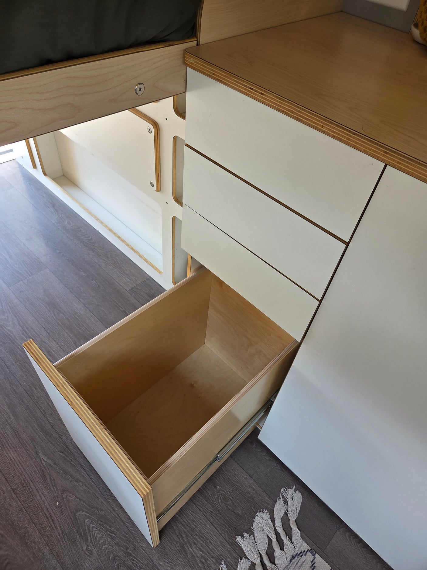 Inside an RV rental, a neatly opened plywood drawer reveals clever storage design amidst white cabinets and dark flooring.