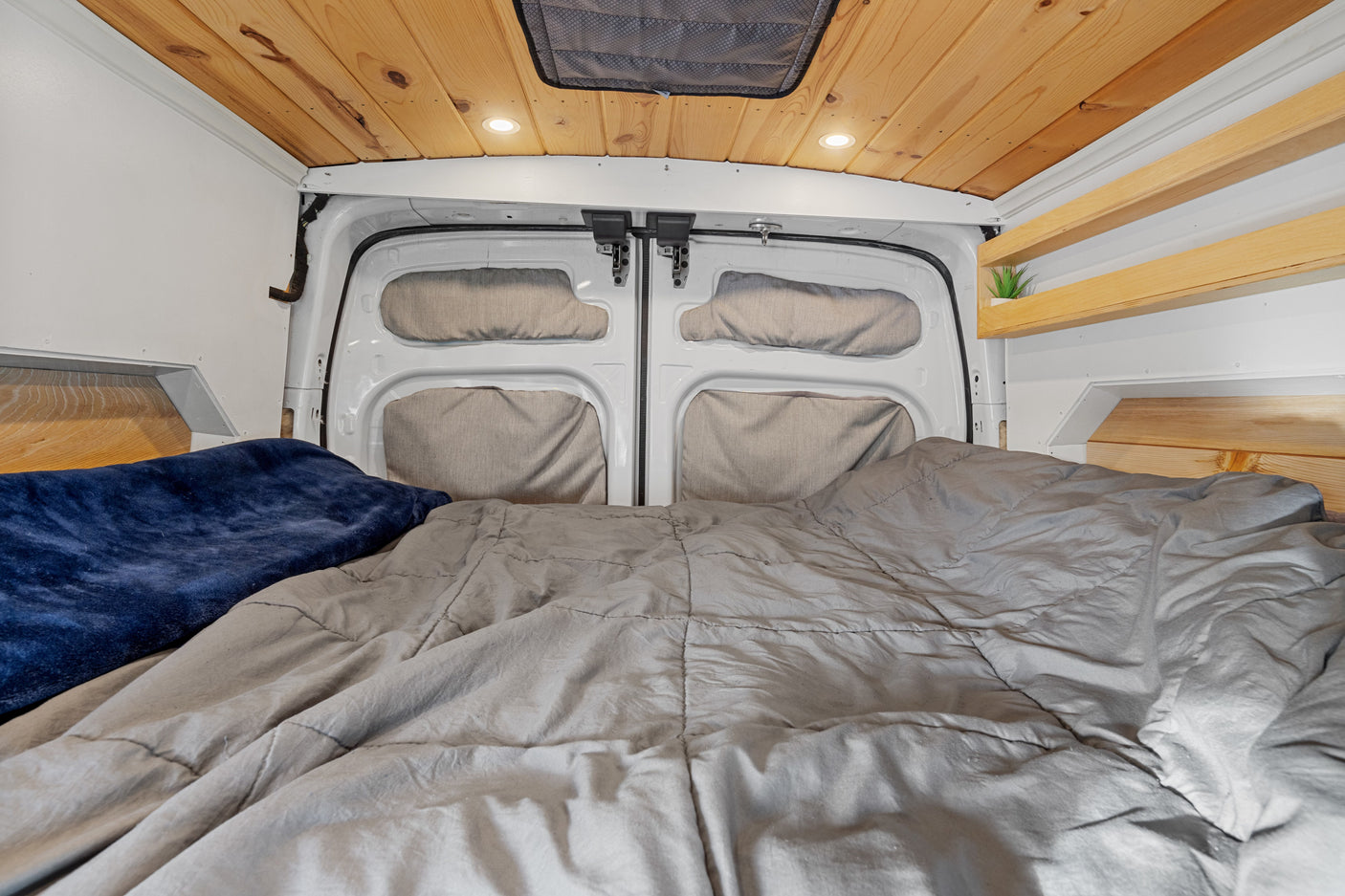 Cozy RV rental bed area with gray comforter, blue blanket, wooden plank ceiling, recessed lights, and rear van doors.