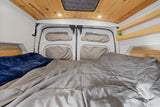 Cozy RV rental bed area with gray comforter, blue blanket, wooden plank ceiling, recessed lights, and rear van doors.