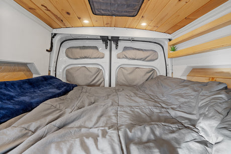 Cozy RV rental bed area with gray comforter, blue blanket, wooden plank ceiling, recessed lights, and rear van doors.